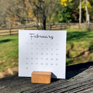 May include: A square white paper calendar page for the month of February with black text and a grid pattern, held upright by a small rectangular wooden block stand. The calendar displays the days of the month from 1 to 28.