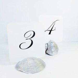 May include: Two white square table number cards featuring the black printed numbers 3 and 4. Each card is held upright by a silver-painted seashell base. The shells have a textured, ridged surface and a metallic finish, providing a stable stand.