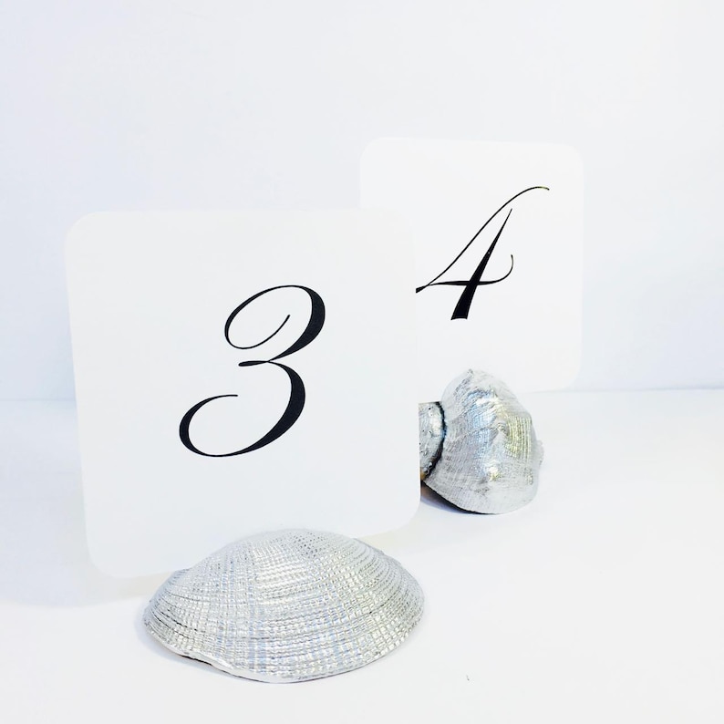 May include: Two white square table number cards featuring the black printed numbers 3 and 4. Each card is held upright by a silver-painted seashell base. The shells have a textured, ridged surface and a metallic finish, providing a stable stand.