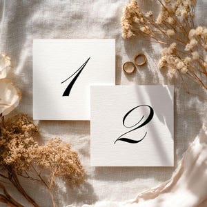 May include: Two square white cards featuring black printed numbers one and two in a calligraphic font. The cards are positioned near two small gold rings and dried beige flowers on a textured off-white fabric surface.