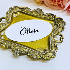 May include: A gold-colored, ornate tray with a white oval card. The card features the name "Olivia" in elegant black script. The tray has a detailed, vintage-style design, suitable for weddings or special events.