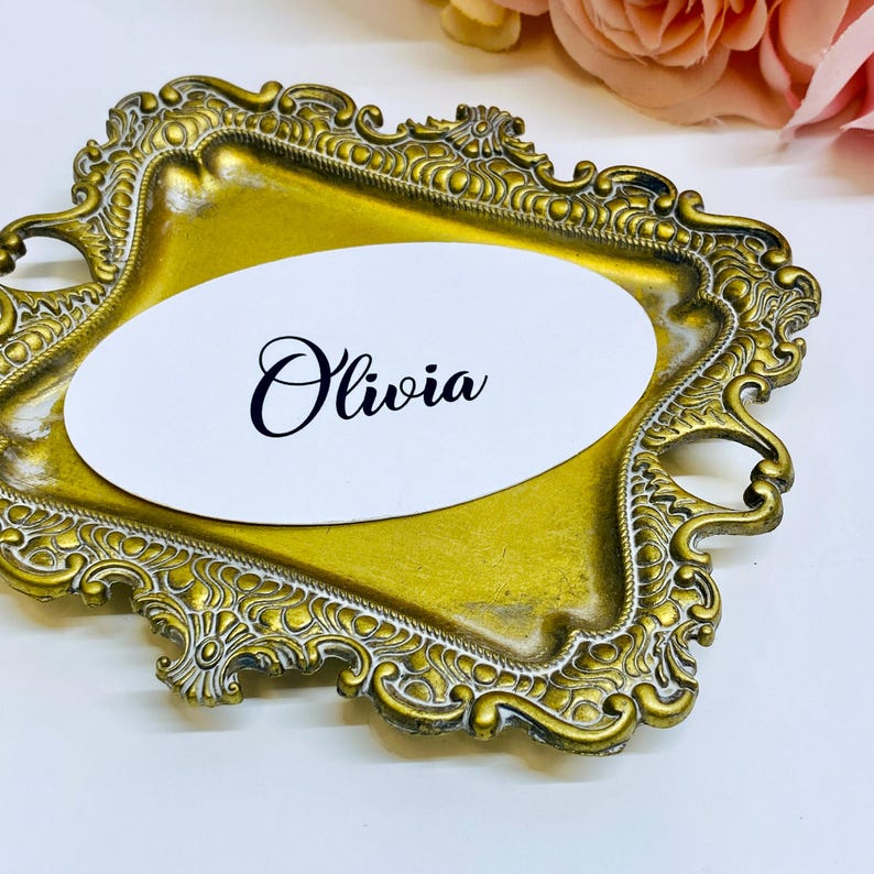 May include: A gold-colored, ornate tray with a white oval card. The card features the name "Olivia" in elegant black script. The tray has a detailed, vintage-style design, suitable for weddings or special events.