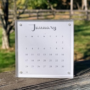 May include: A square acrylic desk calendar featuring the month of January with black printed text and numbers on a white background. The calendar is held together by four small silver-toned metal screws at each corner of the clear frame.