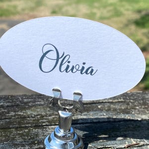 May include: A silver place card holder with a round base and a clip holding a white oval card. The card displays the name "Olivia" in elegant cursive script. The holder sits on a weathered wooden surface.