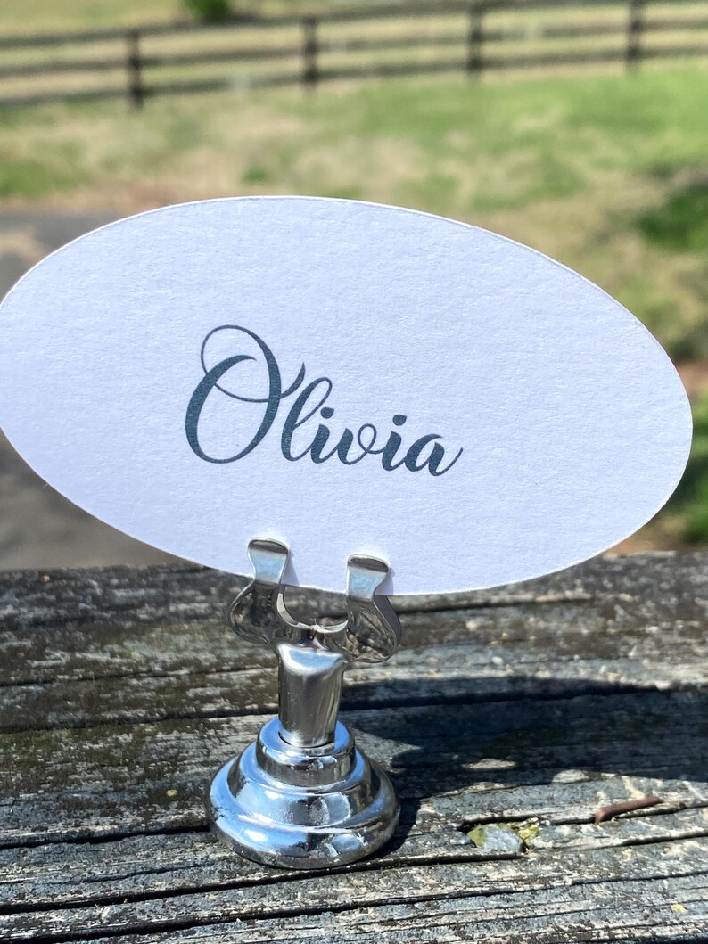 May include: A silver place card holder with a round base and a clip holding a white oval card. The card displays the name "Olivia" in elegant cursive script. The holder sits on a weathered wooden surface.