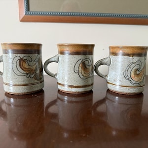 May include: Three ceramic mugs with a speckled, light gray base, brown rims, and handles. Each mug features a brown and orange swirl design. The mugs are displayed on a dark brown surface, with a framed picture in the background.