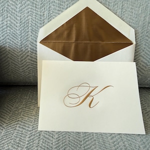 May include: Ivory stationery set featuring a card with a gold script letter "K" and a matching envelope with a gold interior. The set is presented on a light blue patterned surface.