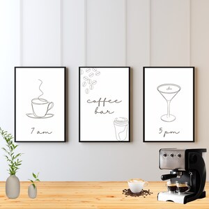 May include: Three framed art prints with black frames. The prints feature line drawings of a coffee cup, coffee beans and a to-go cup, and a martini glass. Text on the prints reads "7 am", "coffee bar", and "5 pm". A coffee machine and coffee cup are on the wooden surface.
