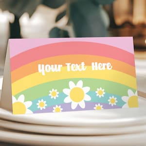 Pode incluir: Um cartão de lugar com arco-íris com um arco-íris rosa, laranja, amarelo, verde, azul e roxo com margaridas brancas. O texto "Your Text Here" está no centro do arco-íris.