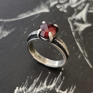 May include: A silver ring with a faceted, deep red gemstone. The ring has a textured band with a dark, oxidised finish, and the gemstone is held by four prongs. The ring is set against a dark background.