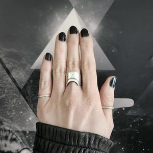 May include: A silver ring featuring a crescent moon design. The ring is worn on the middle finger of a hand with black nail polish.
