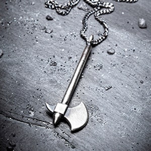 May include: A silver-toned axe pendant necklace. The axe charm has a long handle and a double-bladed head. The necklace chain is a linked design. The background is a textured, grey surface with small dark particles scattered around.