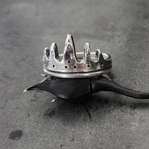 May include: A silver ring featuring a crown-like design with jagged, pointed peaks and a series of small, circular indentations along the base. The metal has a textured, weathered finish, giving it a rugged appearance with dark oxidation in the crevices.