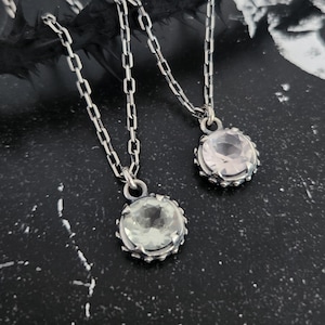 May include: Two silver necklaces featuring round, faceted gemstones set in prong settings. One pendant holds a pale green stone, while the other contains a clear, light-reflecting stone. Both pendants hang from matching silver link chains.