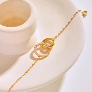 Könnte beinhalten: Ein goldenes Kettenarmband mit zwei ineinandergreifenden Kreisen, die mit klaren Zirkonia-Steinen besetzt sind.