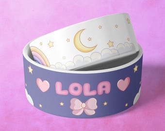 May include: A blue and white ceramic pet bowl with a moon, stars, and clouds design. The bowl has the name "LOLA" written on it in pink letters with a pink bow.