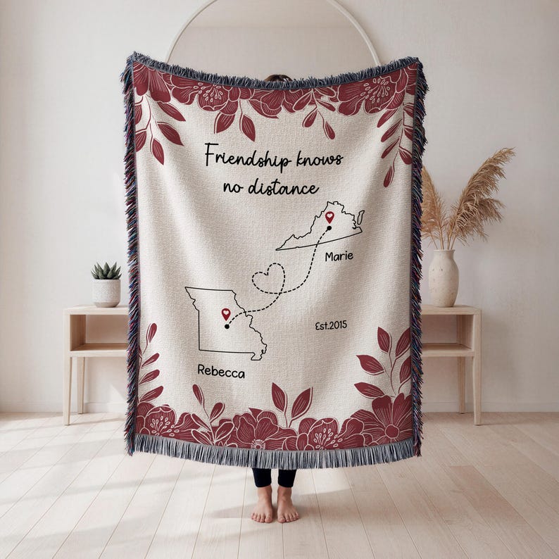 Puede incluir: Una manta tejida beige con un borde y acentos florales burdeos. La manta presenta el texto "Friendship knows no distance" y contornos de dos estados conectados por una l&iacute;nea punteada, con nombres y una fecha.