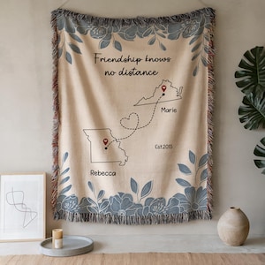 Puede incluir: Manta tejida beige con un borde floral y el texto "Friendship knows no distance". Presenta un dise&ntilde;o de mapa con dos estados conectados por una l&iacute;nea de puntos y los nombres "Rebecca" y "Marie". La manta est&aacute; deshilachada.