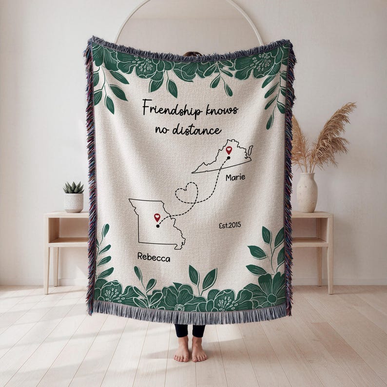 Puede incluir: Una manta tejida de color crema con un borde floral verde oscuro y flecos. La manta presenta el texto "Friendship knows no distance" y contornos de dos estados conectados por una l&iacute;nea punteada, con nombres y una fecha.