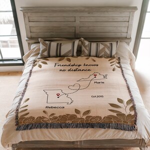 Puede incluir: Una manta beige con un borde floral y el texto "Friendship knows no distance". La manta presenta un mapa con dos estados conectados por una l&iacute;nea de puntos, con nombres y el a&ntilde;o "Est.2015".
