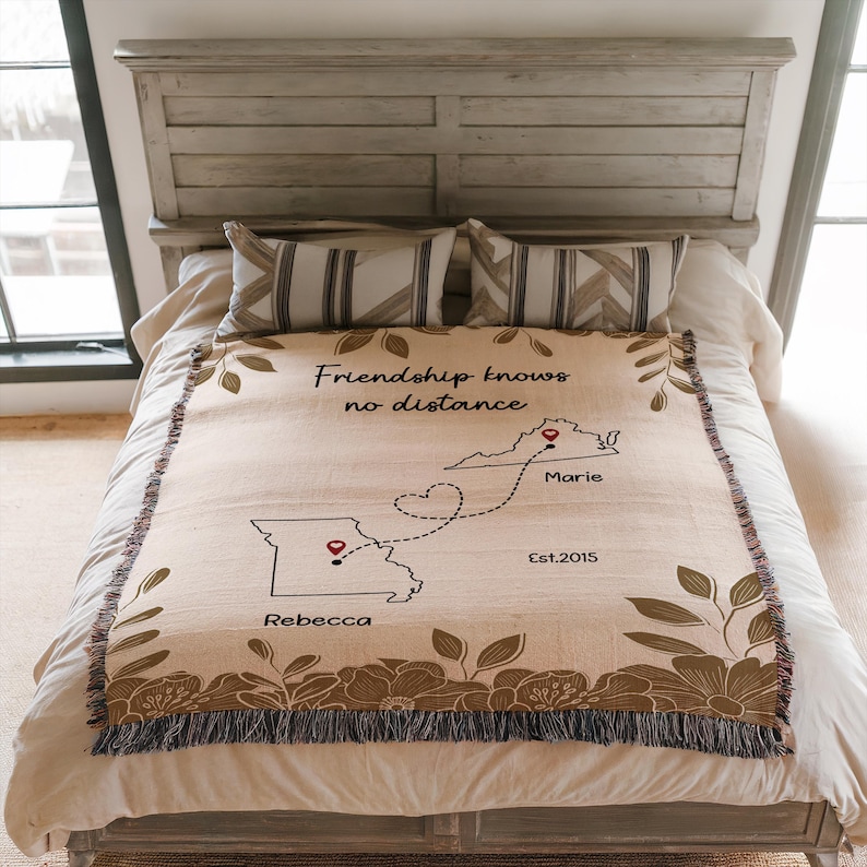 Puede incluir: Una manta beige con un borde floral y el texto "Friendship knows no distance". La manta presenta un mapa con dos estados conectados por una l&iacute;nea de puntos, con nombres y el a&ntilde;o "Est.2015".