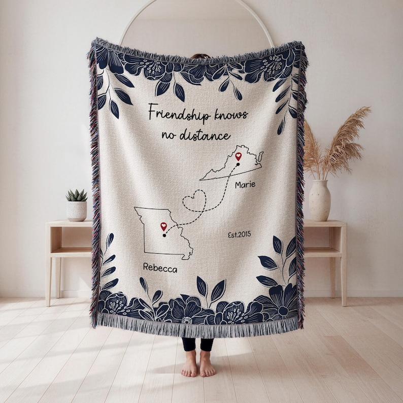 Puede incluir: Manta tejida de color crema con borde floral y de hojas azul marino. La manta presenta el texto "Friendship knows no distance" y contornos de dos estados con marcadores y nombres. La manta mide 152 cm por 127 cm.