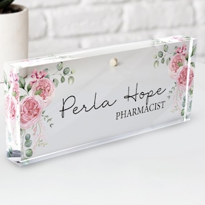 May include: A clear acrylic desk nameplate with a floral design featuring pink roses and green leaves. The nameplate reads "Perla Hope Pharmacist".
