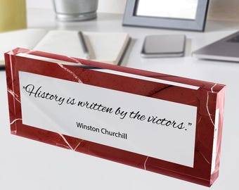 May include: A rectangular, red marble-effect desk block with the quote "History is written by the victors." by Winston Churchill. The block is on a white desk with a yellow mug, a potted plant, and a laptop in the background.