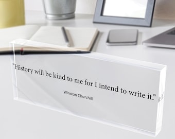 May include: A clear acrylic block with the quote "History will be kind to me for I intend to write it." by Winston Churchill. The block sits on a white desk with a yellow mug, a potted plant, and a picture frame.