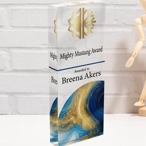 May include: Clear acrylic award with gold accents and the text "Mighty Mustang Award" and "Awarded to Breena Akers." The bottom of the award features a blue and gold abstract design. A wooden poseable figure is in the background.