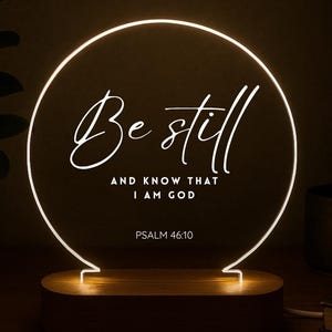 May include: A glowing LED night light with a wooden base. The light features a circular design with the words "Be still and know that I am God" and Psalm 46:10 in white script. The light is on a wooden surface.