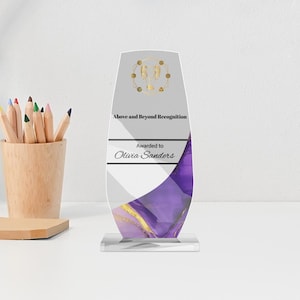 May include: A clear acrylic award with a white and purple marbled design, gold accents, and the text "Above and Beyond Recognition." The award is inscribed "Awarded to Olivia Sanders."