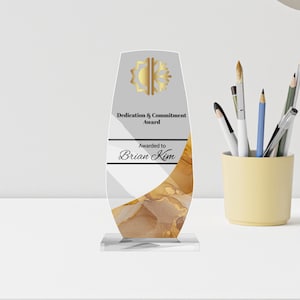 May include: A clear acrylic award with a gold and white design. The award is inscribed with "Dedication & Commitment Award" and "Awarded to Brian Kim". The award is on a clear base and has a gold sunburst design at the top.