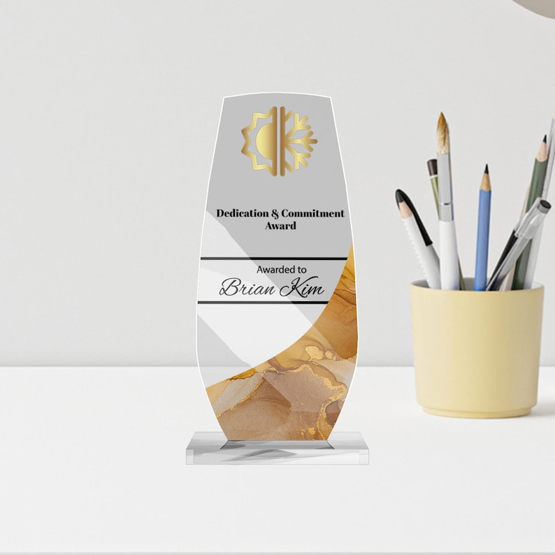 May include: A clear acrylic award with a gold and white design. The award is inscribed with "Dedication & Commitment Award" and "Awarded to Brian Kim". The award is on a clear base and has a gold sunburst design at the top.