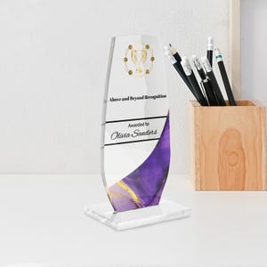 May include: A clear acrylic award with a curved design. The award features the text "Above and Beyond Recognition" and "Awarded to Olivia Sanders." The design includes purple and gold accents.