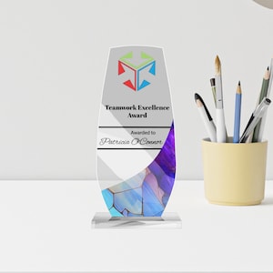 May include: A clear acrylic Teamwork Excellence Award with a colorful geometric design. The award features a logo with red, green, and blue shapes, and text that reads "Awarded to Patricia O'Connor."