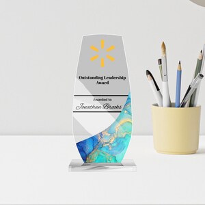 May include: An award for outstanding leadership. The clear acrylic award has a gray top with a yellow starburst logo and text. The bottom has a blue and gold marbled design. The award is on a clear base and the text reads "Awarded to Jonathan Brooks."