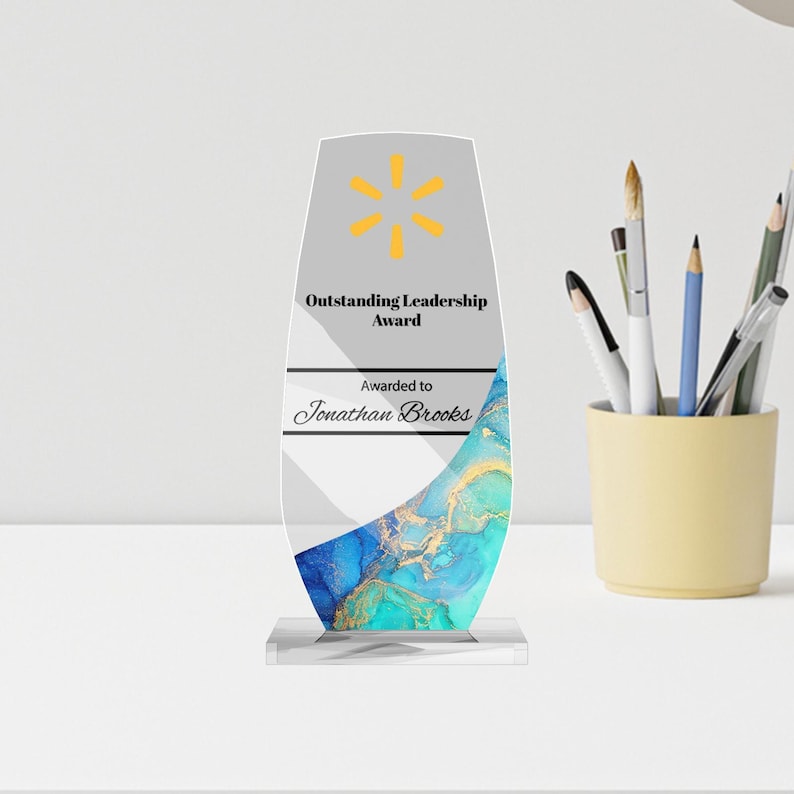 May include: An award for outstanding leadership. The clear acrylic award has a gray top with a yellow starburst logo and text. The bottom has a blue and gold marbled design. The award is on a clear base and the text reads "Awarded to Jonathan Brooks."