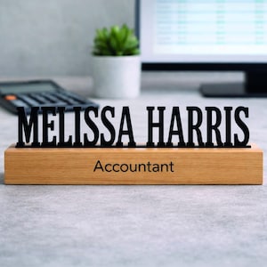 May include: A wooden desk nameplate with the name "MELISSA HARRIS" in black letters. Below the name, the word "Accountant" is printed in black. The nameplate is on a desk with a calculator and a small potted plant.