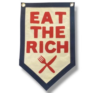 May include: A cream-colored fabric wall banner with a pointed bottom edge and dark blue borders. The text EAT THE RICH is printed in bold red capital letters, followed by a red graphic of a crossed fork and knife. Two metal grommets are at the top.