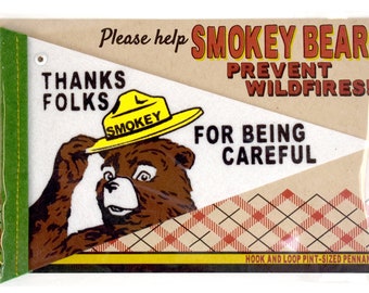 Può includere: Un gagliardetto con Smokey Bear con un cappello giallo, con il testo "Please help Smokey Bear prevent wildfires! Thanks folks for being careful." Il gagliardetto ha un bordo verde e un motivo a quadri.