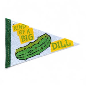 May include: A white pennant with a green border and a green pickle illustration. The pennant has the words "KIND OF A BIG" and "DILL" in yellow block letters. A fun, food-themed item.