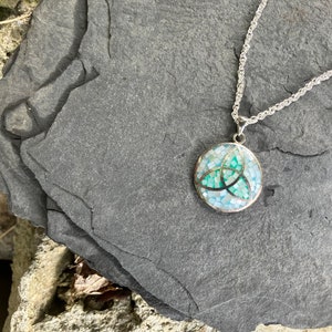May include: A silver chain necklace with a round pendant featuring a green and blue mosaic Celtic knot design.