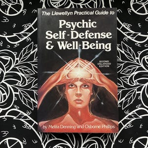 May include: A book titled "The Llewellyn Practical Guide to Psychic Self-Defense & Well-Being" with a woman's face on the cover. The cover art features a woman with hands raised to her head, with text above and below the image. The background is black and white.