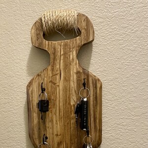 May include: Wooden key holder with a rope handle and five hooks. The holder is mounted on a wall and has several keys hanging from it.