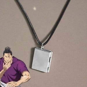 May include: A silver-colored rectangular pendant necklace with a black cord. The pendant has a hinged design, suggesting it may open. The background is a solid, neutral color.