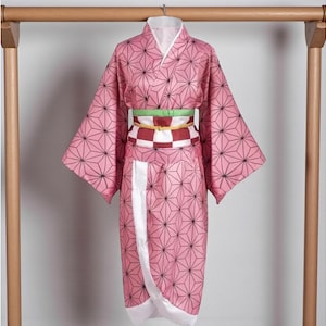 May include: Pink kimono with a geometric pattern of black lines and shapes. The garment has wide sleeves and a white collar. A green and red checkered belt is wrapped around the waist. The kimono is hanging on a wooden rack.