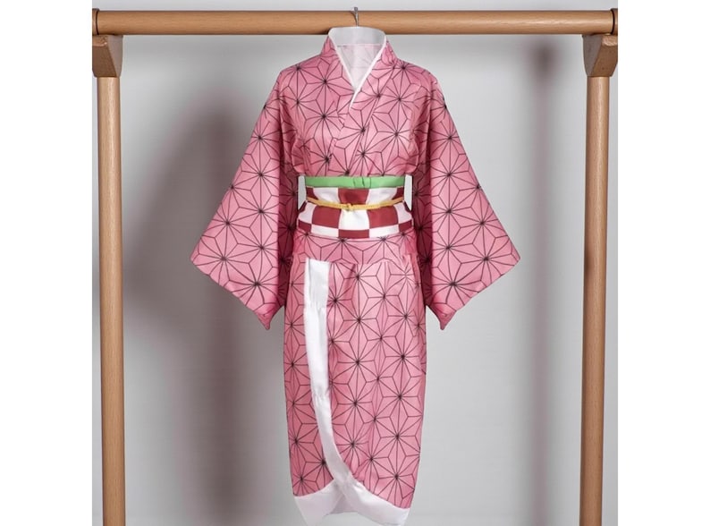 May include: Pink kimono with a geometric pattern of black lines and shapes. The garment has wide sleeves and a white collar. A green and red checkered belt is wrapped around the waist. The kimono is hanging on a wooden rack.