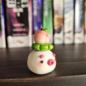 May include: A white ceramic figurine with a pink peach on top, green leaves, and a pink and brown face. The figurine is sitting on a wooden shelf.
