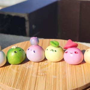 May include: Six small, round clay figures in various colours, including white, green, pink, and yellow. Each figure has a unique design, some with facial features. They are displayed on a wooden surface, with a dark background.